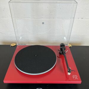 Red turntable with a clear dust cover, black platter mats, and a tonearm on the right, set on a black table for display.